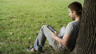 Ein Mann sitzt an einen Baumstamm lehnend auf einer Wiese und liest ein E-Book.