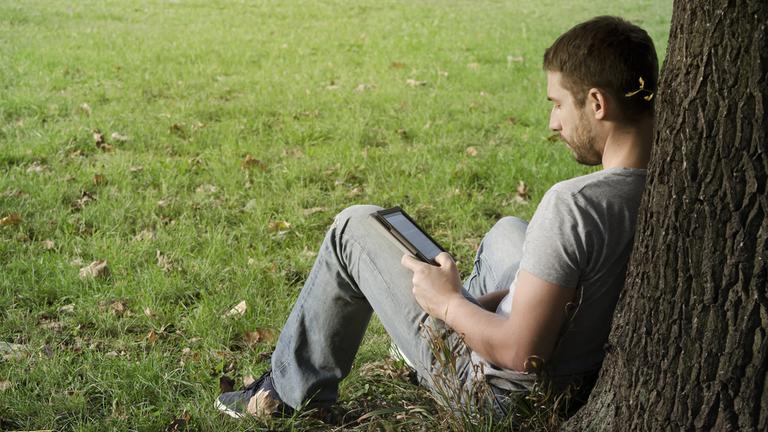 Ein Mann sitzt an einen Baumstamm lehnend auf einer Wiese und liest ein E-Book.