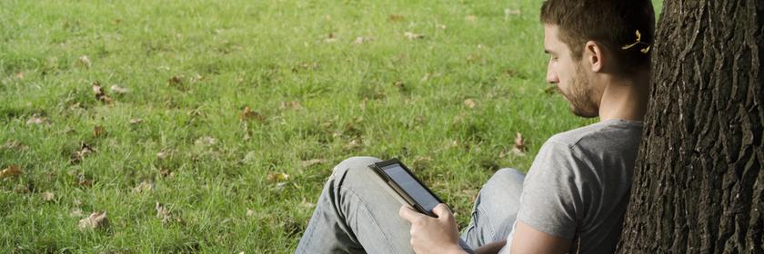 Ein Mann sitzt an einen Baumstamm lehnend auf einer Wiese und liest ein E-Book.