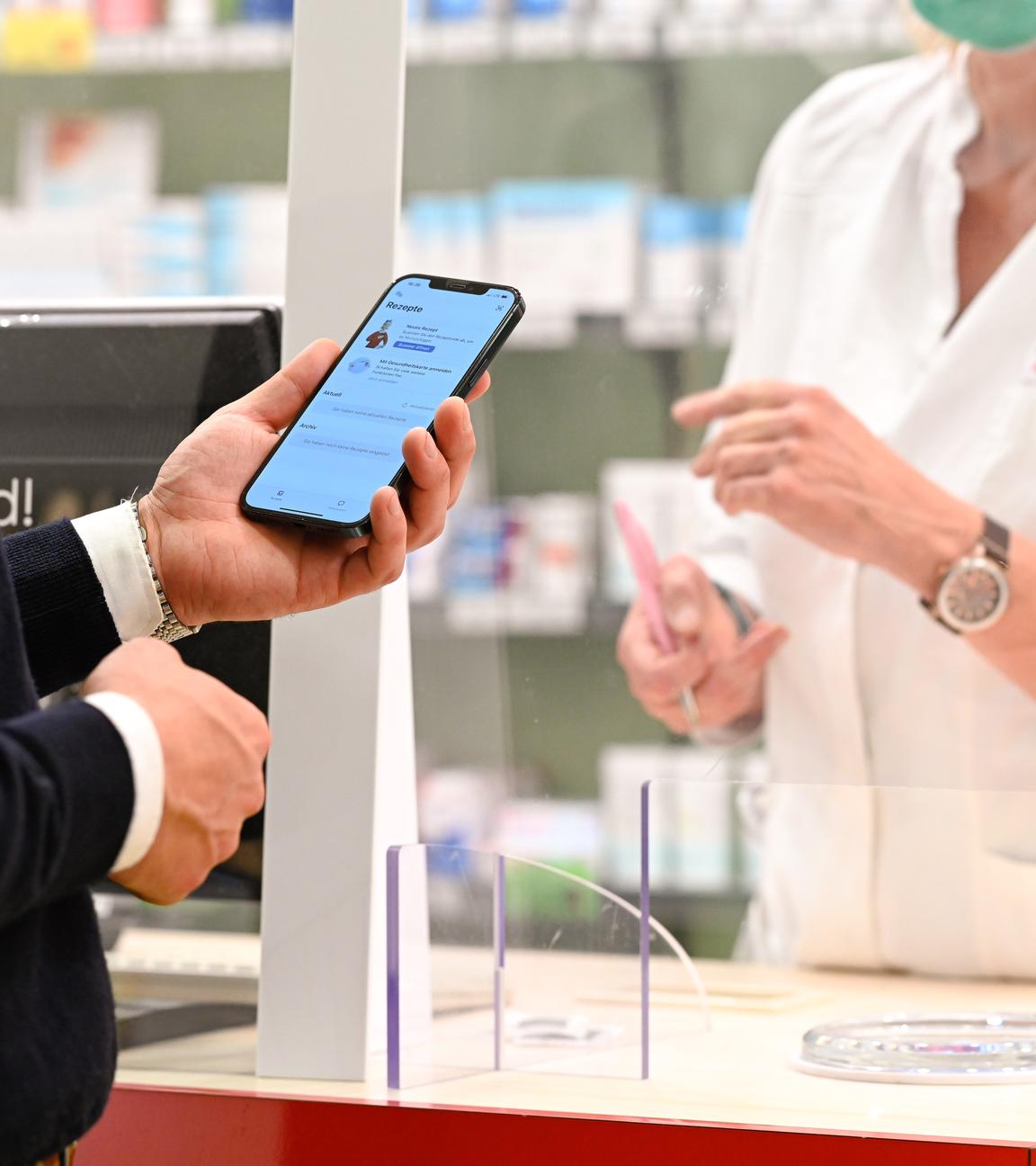 Ein Mann mit seinem Handy in der Hand, auf dem die App «Das E-Rezept» geöffnet ist, wird in einer Apotheke von einer Apothekerin bedient