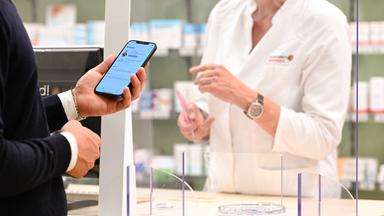 Ein Mann mit seinem Handy in der Hand, auf dem die App «Das E-Rezept» geöffnet ist, wird in einer Apotheke von einer Apothekerin bedient