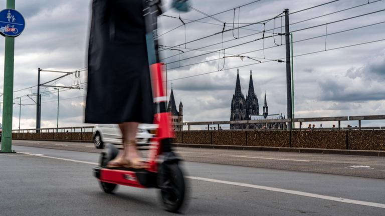 Frau fährt auf einem E-Scooter über die Deutzer Brücke in Köln, im Hintergrund der Kölner Dom.