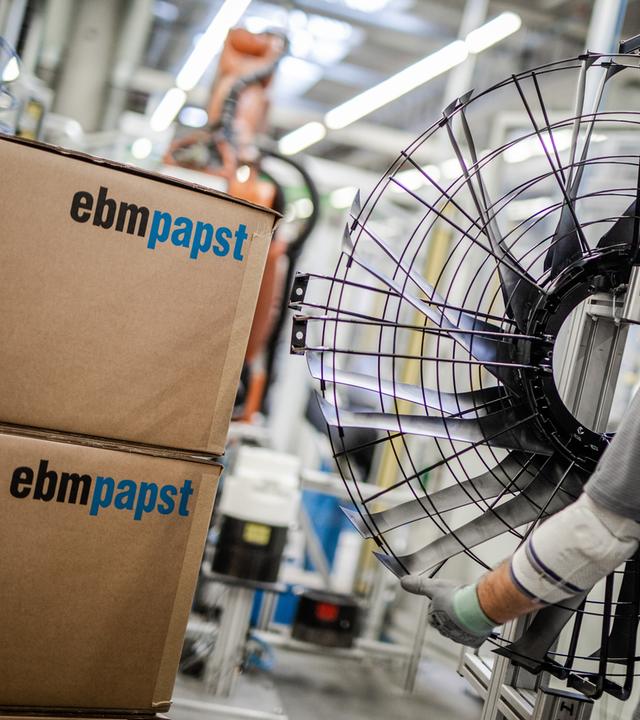 Ein Mitarbeiter des Elektromotoren- und Ventilatorenherstellers EBM-Papst arbeitet in der Produktion an einem Ventilator. Neben ihm stehen zwei aufeinander gestapelte EBM-Papst Pakete.
