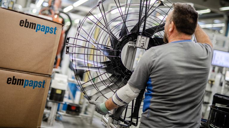 Ein Mitarbeiter des Elektromotoren- und Ventilatorenherstellers EBM-Papst arbeitet in der Produktion an einem Ventilator. Neben ihm stehen zwei aufeinander gestapelte EBM-Papst Pakete.