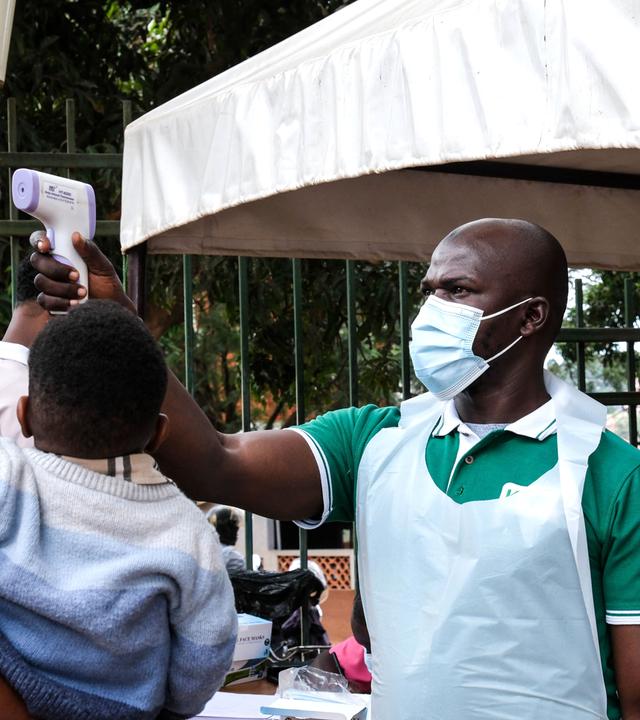 Ein Gesundheitsbeamter untersucht einen Mann mit einem Kind auf dem Arm mit einem Thermometer vor dem Kibuli Muslim Hospital in Kampala.