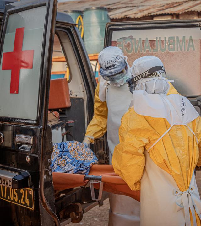 Zwei mit Schutzanzügen bekleidete Personen heben eine Trage in einen Krankenwagen.