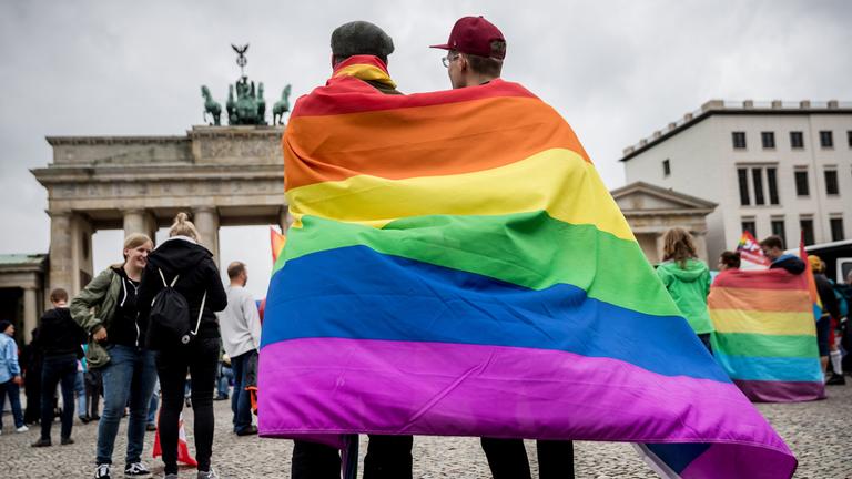 Zwei Männer stehen vor dem Brandenburger Tor in Berlin mit einer Regenbogenflagge.