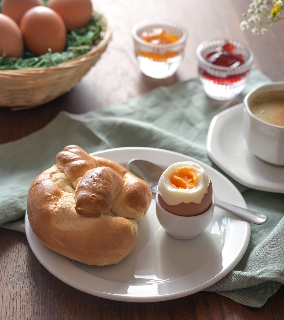 Frühstück mit einem Osternest mit Eiern, Marmelade und Kaffeetasse im Hintergrund sowie ein Teller mit einem Hefebrötchen und einem gekochten Ei in einem Eierbecher. 