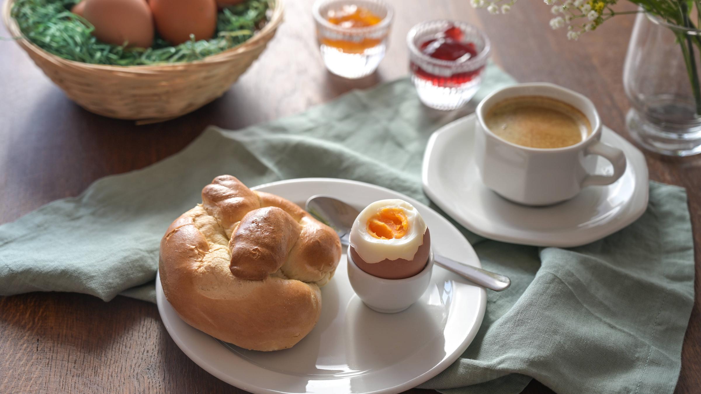 Frühstück mit einem Osternest mit Eiern, Marmelade und Kaffeetasse im Hintergrund sowie ein Teller mit einem Hefebrötchen und einem gekochten Ei in einem Eierbecher. 