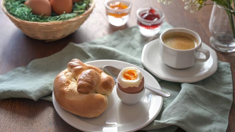 Frühstück mit einem Osternest mit Eiern, Marmelade und Kaffeetasse im Hintergrund sowie ein Teller mit einem Hefebrötchen und einem gekochten Ei in einem Eierbecher. 