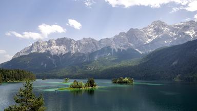 Die Sonne scheint bei blauem Himmel und wenigen Wolken auf die Landschaft am Eibsee, im Hintergrund ist das Zugspitzmassiv zu sehe