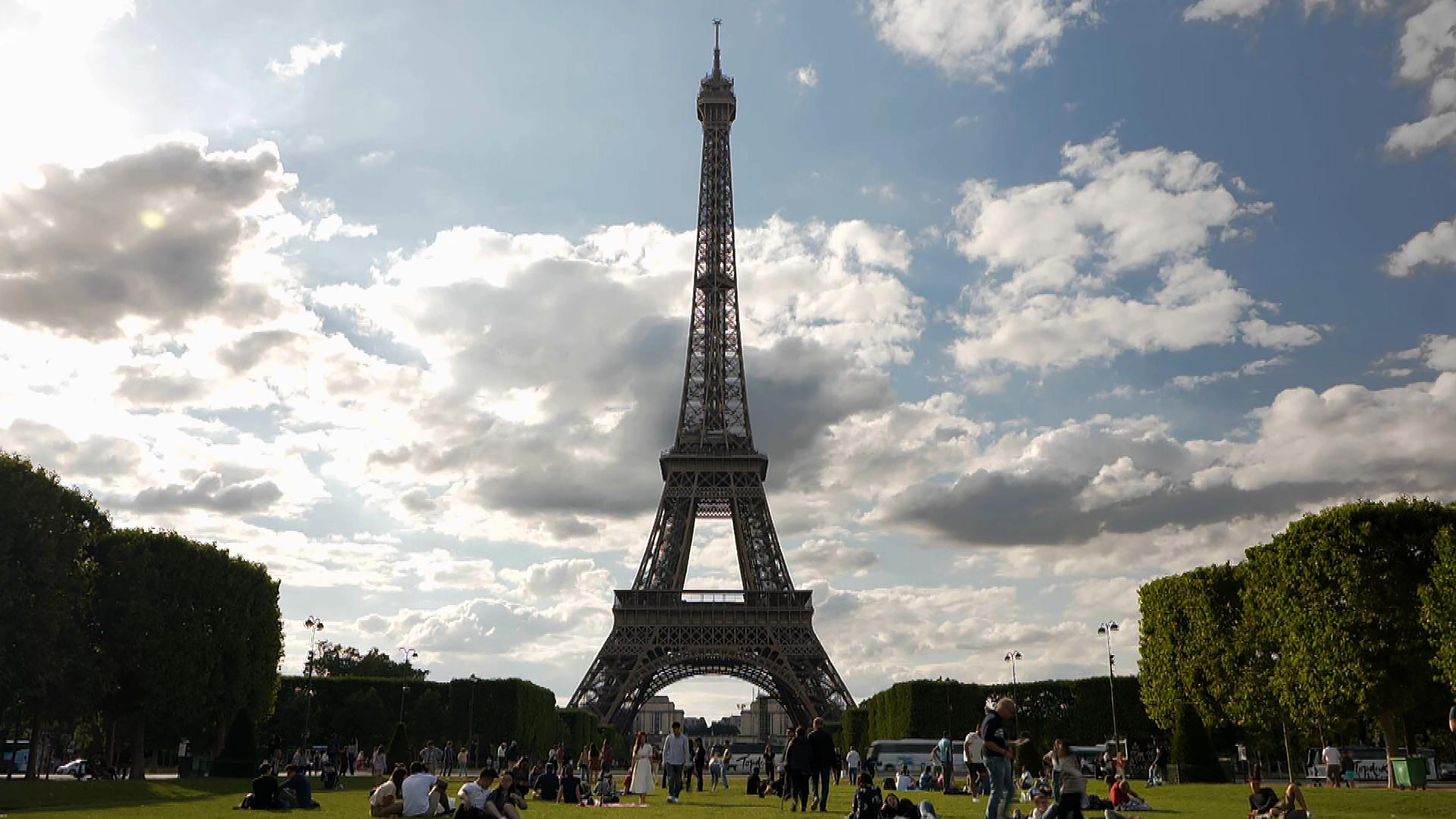 "Eiffels Superbauten - Giganten aus Stahl und Eisen": Der Eiffelturm in der Totalen, davor Menschen auf einer Wiese, am Rand säumen Bäume das Bild, Wolken am Himmel.
