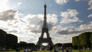 "Eiffels Superbauten - Giganten aus Stahl und Eisen": Der Eiffelturm in der Totalen, davor Menschen auf einer Wiese, am Rand säumen Bäume das Bild, Wolken am Himmel.