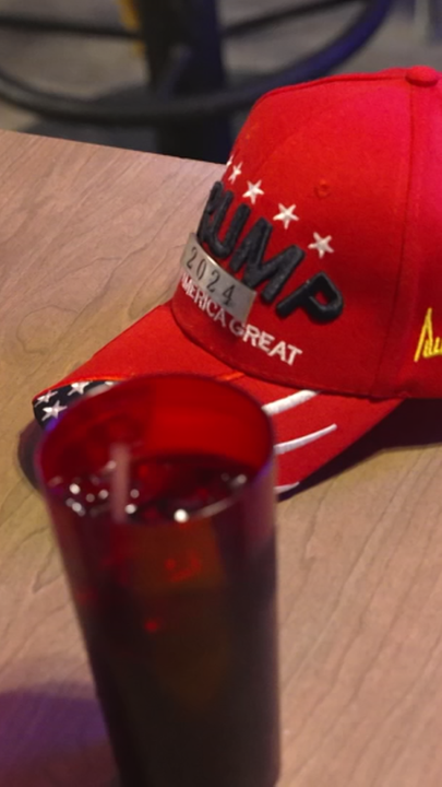 Eine Rote Mütze mit der Aufschrift Trump 2024 liegt auf einem Tisch, davor steht ein Glas mit einem Getränk.