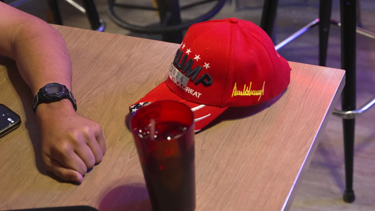 Eine Rote Mütze mit der Aufschrift Trump 2024 liegt auf einem Tisch, davor steht ein Glas mit einem Getränk.