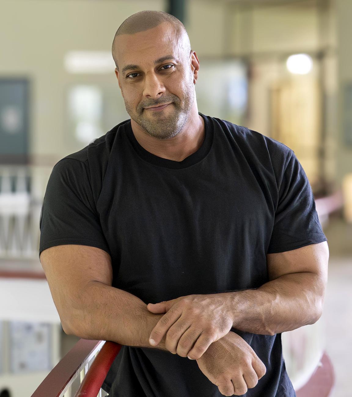 Lehrer Reff lehnt am Treppengeländer in seiner Schule. Auf der Treppe sind mehrere Schüler zu sehen.