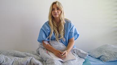 Eine Frau im Kittel sitzt auf einem Krankenhausbett, über ihren Beinen hat sie eine Decke.