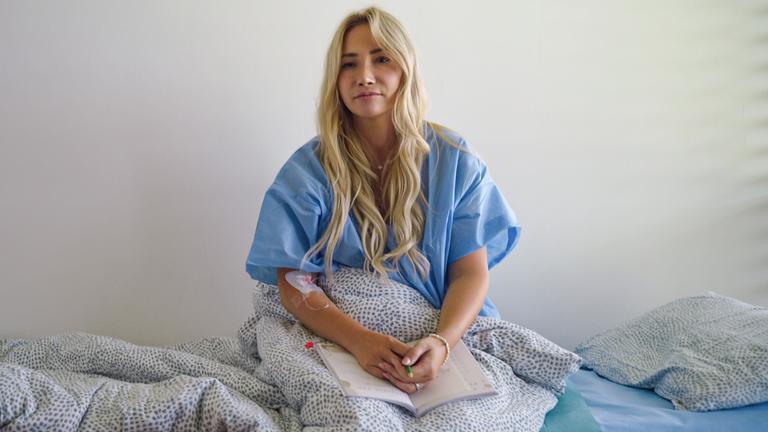 Eine Frau im Kittel sitzt auf einem Krankenhausbett, über ihren Beinen hat sie eine Decke.