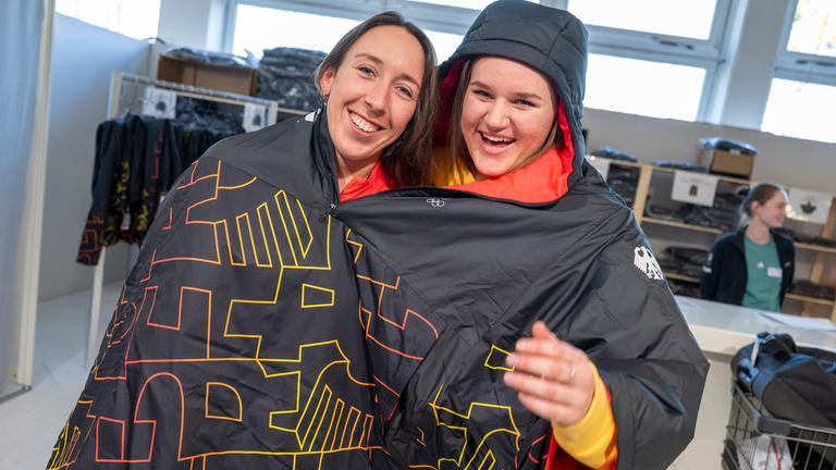Kira Weidle-Winkelmann (l.) und Emma Aicher teilen sich während der Einkleidung für die Winterspiele in Mailand aus Spaß einen Poncho