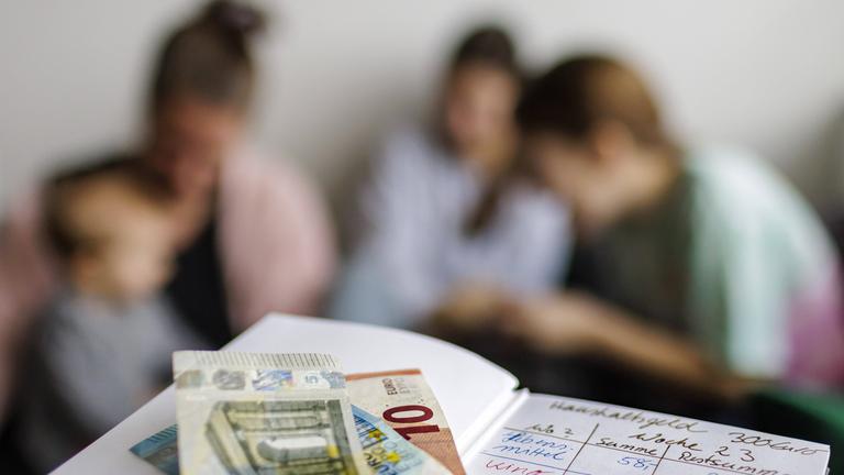 Symbolbild: Haushaltsbuch mit fuenfunddreissig Euro. Im Hintergrund eine Mutter mit drei Kindern. 