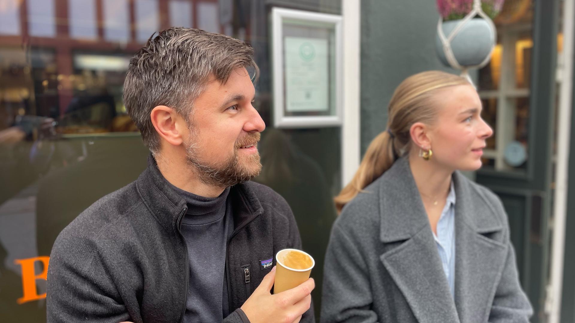 Jakob Kristensen und Anna Horst sitzen vor einem Coffee-Shop und schauen zur Seite.