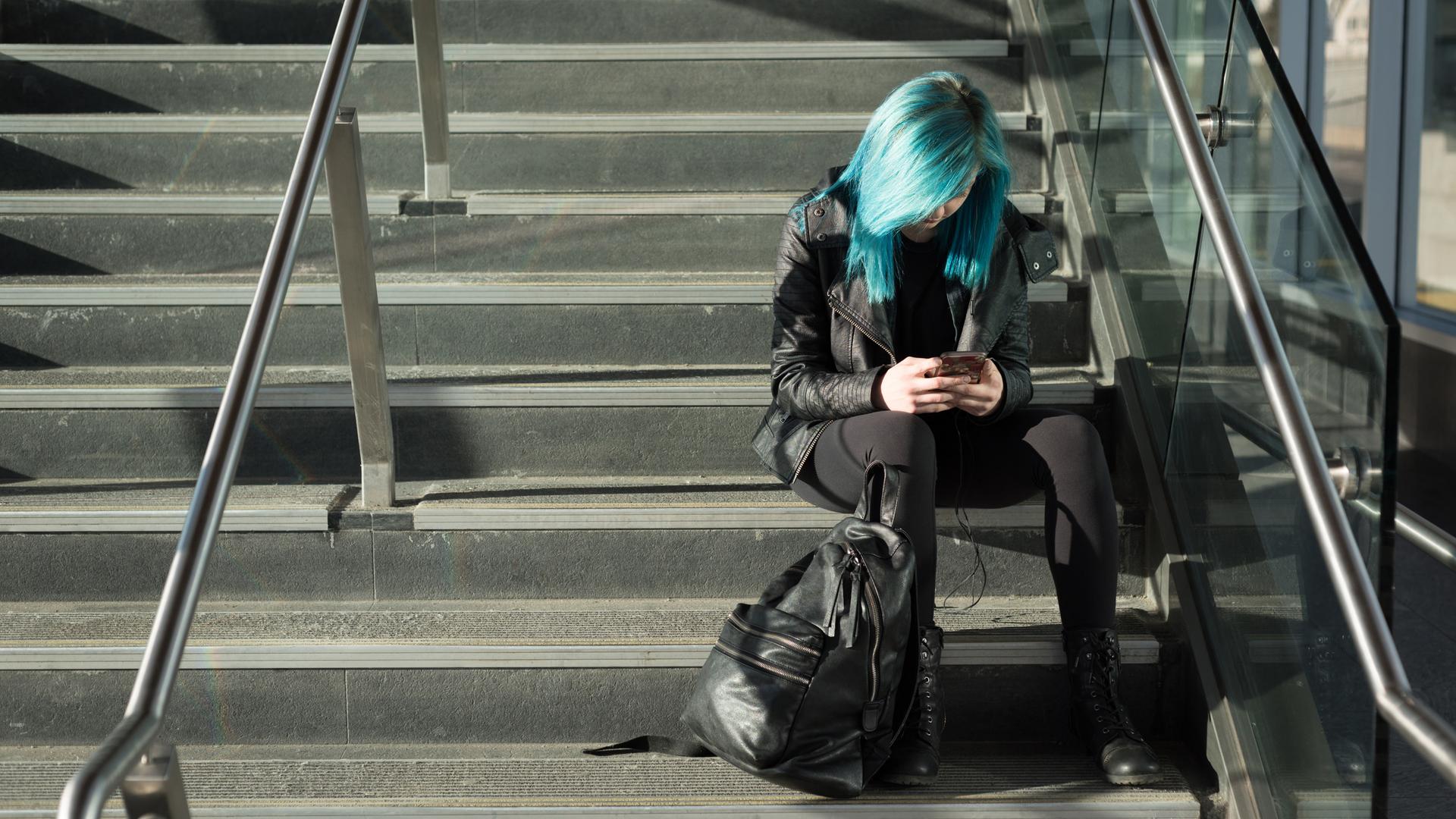 Blauhaarige Jugendliche sitzt alleine auf ein Treppe und schaut auf ihr Smartphone.