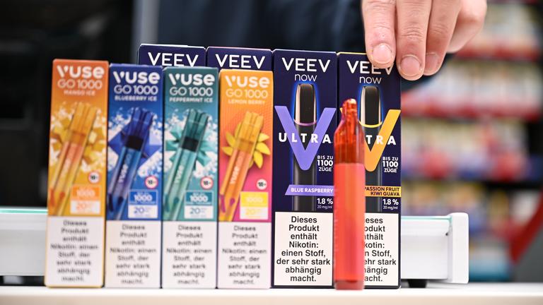 Mehrere Packungen mit Einweg-E-Zigaretten verschiedener Marken nebeneinander aufgestellt, davor steht eine ausgepackte Einweg-E-Zigarette.