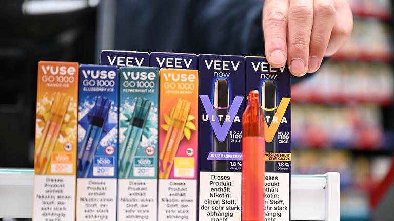 Eine Hand greift nach einer Packung Einweg-E-Zigaretten, die auf der Verkaufstheke eines Tabakladens stehen. 