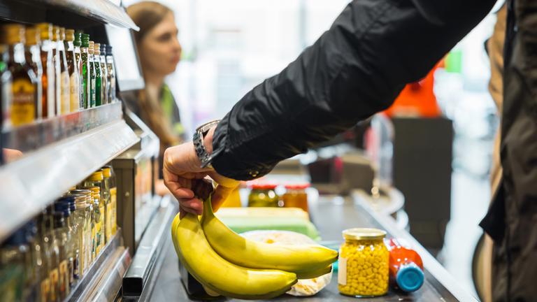 Person legt Bananen zusammen mit anderen Artikeln auf das Förderband an der Supermarktkasse, während im Hintergrund eine Kassiererin zu sehen ist.