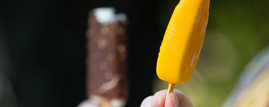Im Vordergrund wird ein orangefarbenes Eis am Stiel in der Hand gehalten, unscharf im Hintergrund eines, das mit Schokolade überzogen ist.