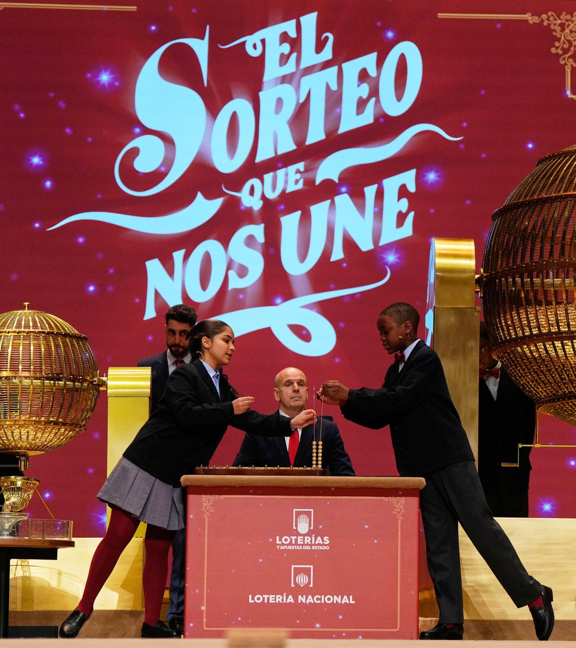 Zwei Kinder ziehen während der jährlichen Weihnachtslotterie El Gordo («Der Dicke») im Madrider Teatro Real die Lottozahlen. Im Hintergrund zwei Männer, dahinter eine rote Wand. Vorne zwei goldene große Gitterkugeln.