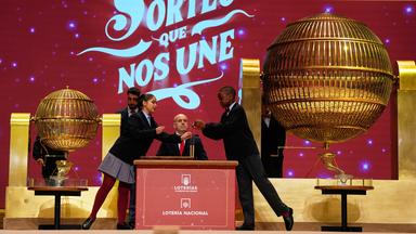 Zwei Kinder ziehen während der jährlichen Weihnachtslotterie El Gordo («Der Dicke») im Madrider Teatro Real die Lottozahlen. Im Hintergrund zwei Männer, dahinter eine rote Wand. Vorne zwei goldene große Gitterkugeln.