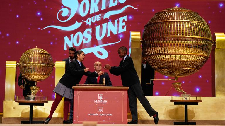 Zwei Kinder ziehen während der jährlichen Weihnachtslotterie El Gordo («Der Dicke») im Madrider Teatro Real die Lottozahlen. Im Hintergrund zwei Männer, dahinter eine rote Wand. Vorne zwei goldene große Gitterkugeln.