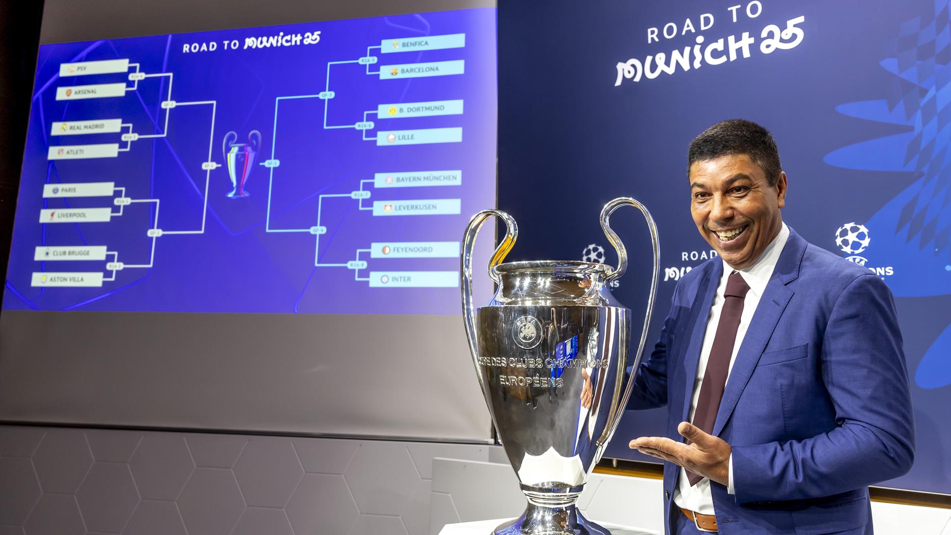 Giovane Elber steht nach der Champions-League-Auslosung neben dem Pokal und lächelnd in die Kamera.
