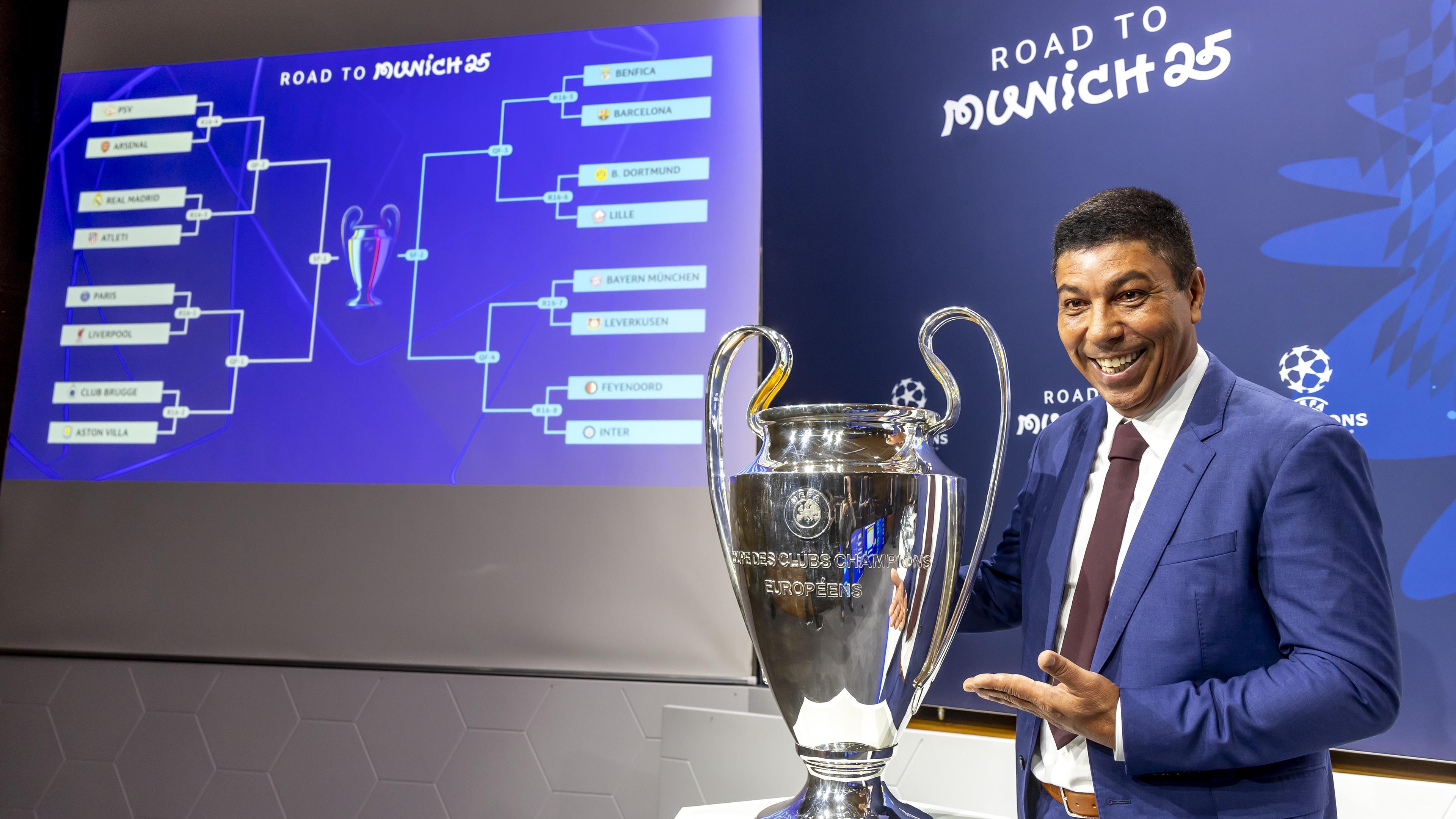 Giovane Elber steht nach der Champions-League-Auslosung neben dem Pokal und lächelnd in die Kamera.