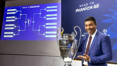 Giovane Elber steht nach der Champions-League-Auslosung neben dem Pokal und lächelnd in die Kamera.