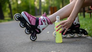 Frau mit Inline-Skates sitzt auf einer Parkbank und greift zur Flasche mit einem Elektrolyt-Drink.