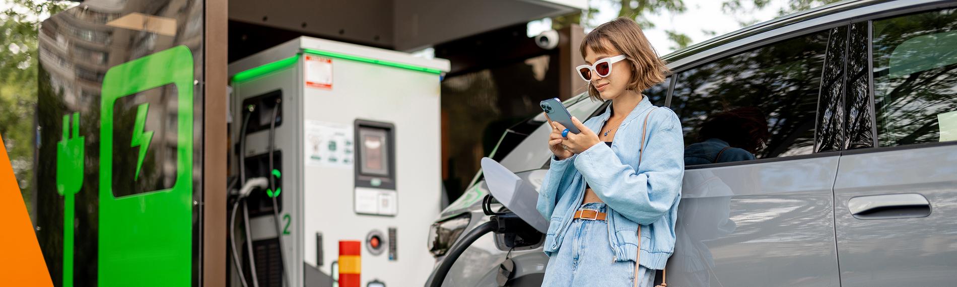 Frau lädt Elektroauto und schaut währendessen in ihr Smartphone