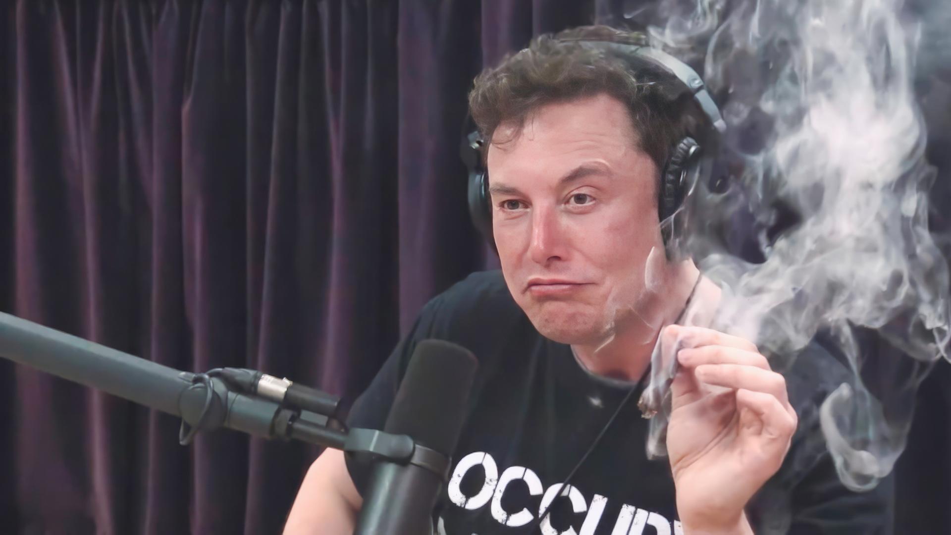 Elon Musk, mit schwarzem T-Shirt mit weißer Aufschrift, sitzt während eines Interviews im Jahr 2018 vor einem Mikrophon und hält in der linken Hand einen Joint, der qualmt.