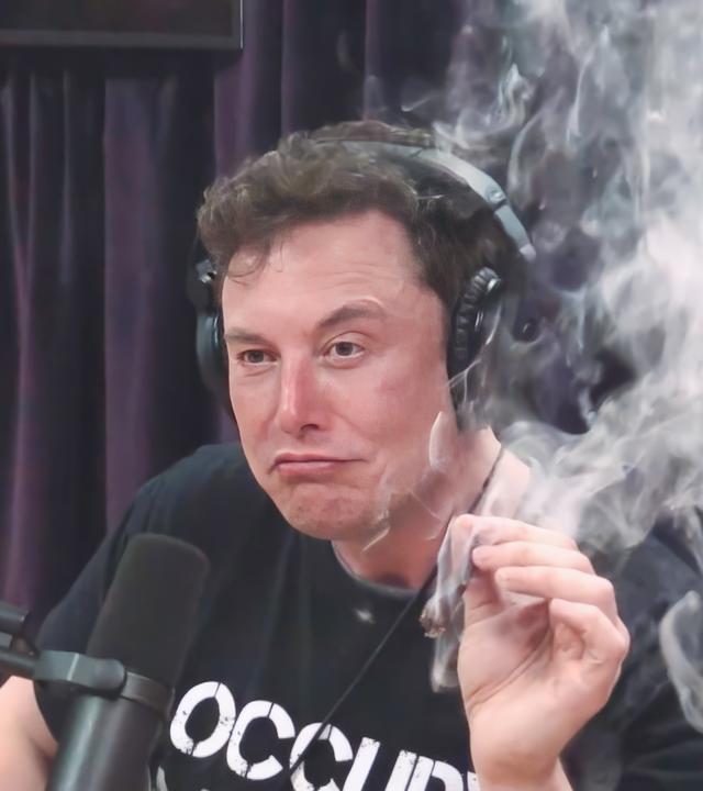 Elon Musk, mit schwarzem T-Shirt mit weißer Aufschrift, sitzt während eines Interviews im Jahr 2018 vor einem Mikrophon und hält in der linken Hand einen Joint, der qualmt.
