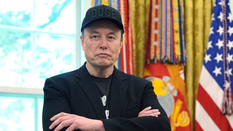 Elon Musk verschränkt die Arme. Im Hintergrund sind Teile des Oval Office des US-Präsidenten Trump zu sehen