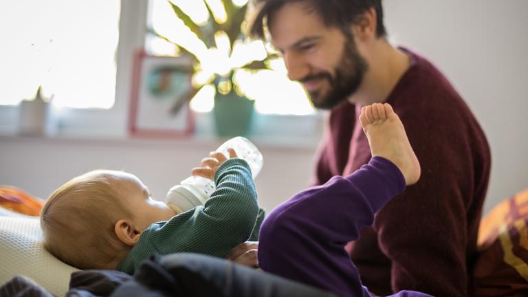Ein Mann schaut auf sein Kind, welches an einer Flasche nuckelt.