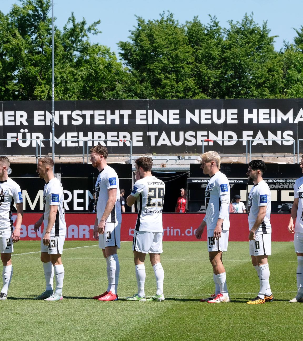 Elversberger Spieler stehen in der Ursapharm-Arena vor einem Plakat, auf dem geschrieben steht, dass das Stadion erweitert wird.