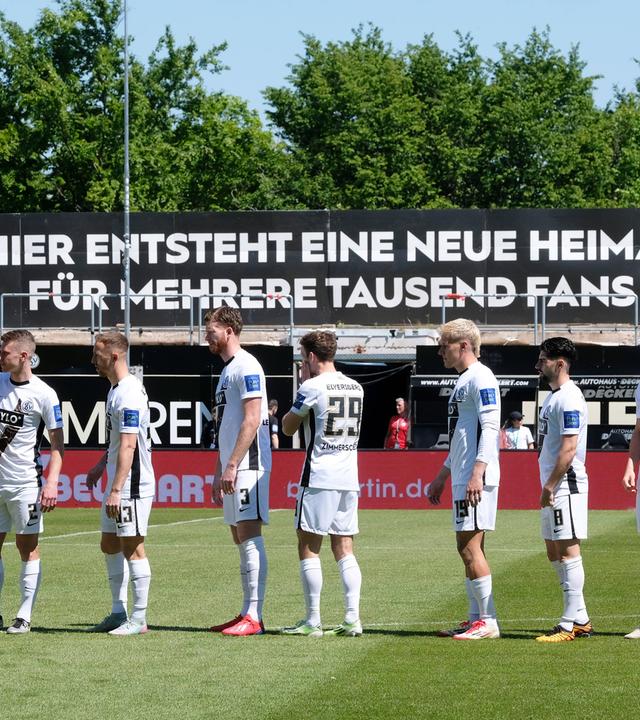 Elversberger Spieler stehen in der Ursapharm-Arena vor einem Plakat, auf dem geschrieben steht, dass das Stadion erweitert wird.
