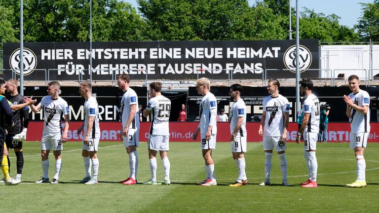 Elversberger Spieler stehen in der Ursapharm-Arena vor einem Plakat, auf dem geschrieben steht, dass das Stadion erweitert wird.