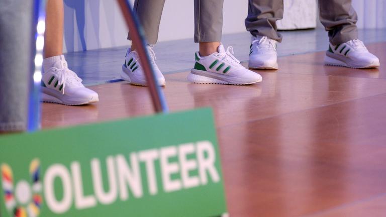 Im Vordergrund ein Schild mit der Aufschrift "Volunteer". Im Hintergrund Beine der ehrenamtlichen Helfer.