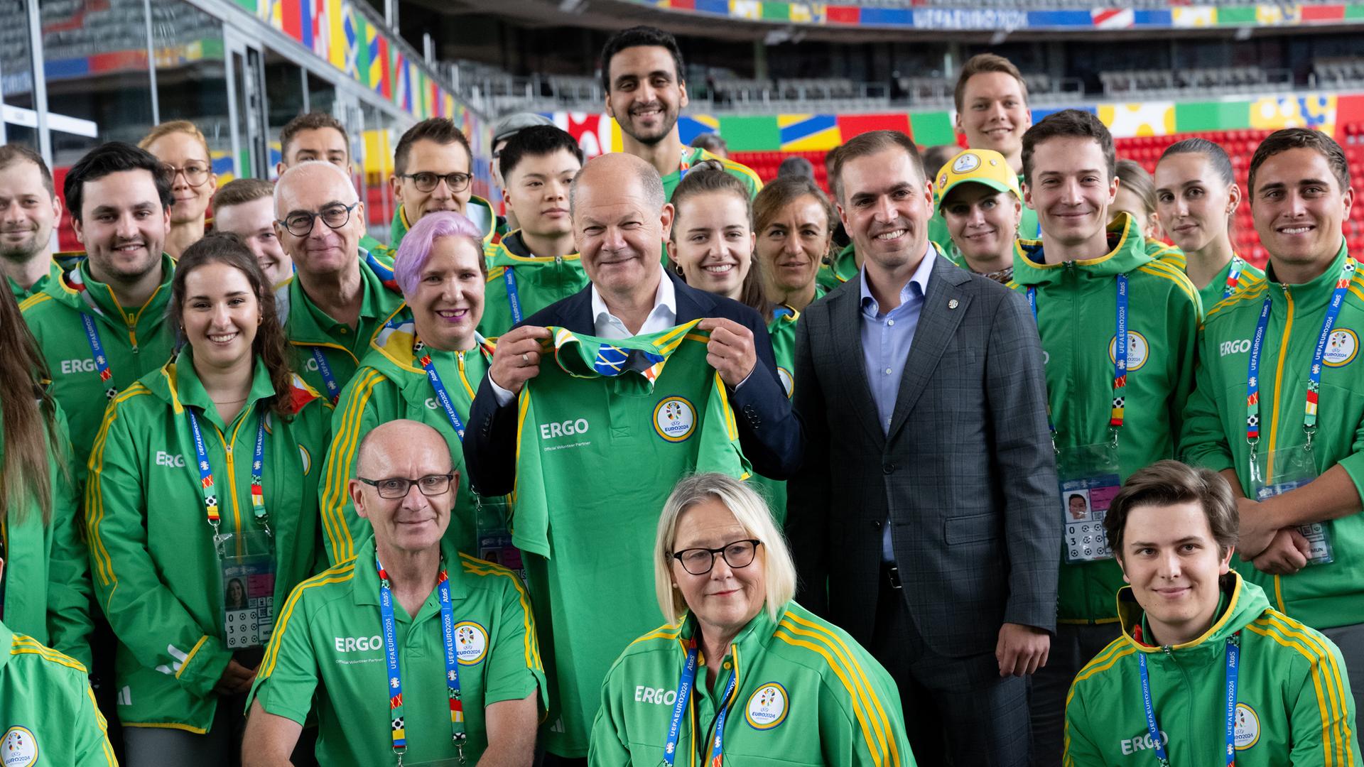 Bundeskanzler Olaf Scholz und Philipp Lahm schauen mit einer Gruppe Volunteers in der München Fußball Arena in die Kamera. Scholz hält ein Trikot mit der Aufschrift "Volunteer" in der Hand.