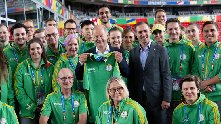 Bundeskanzler Olaf Scholz und Philipp Lahm schauen mit einer Gruppe Volunteers in der München Fußball Arena in die Kamera. Scholz hält ein Trikot mit der Aufschrift "Volunteer" in der Hand.