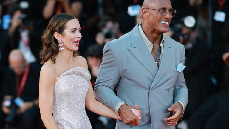 Dwayne Johnson und Emily Blunt auf der Roten Treppe in Venedig. Sie halten sich an der Hand und lächeln.