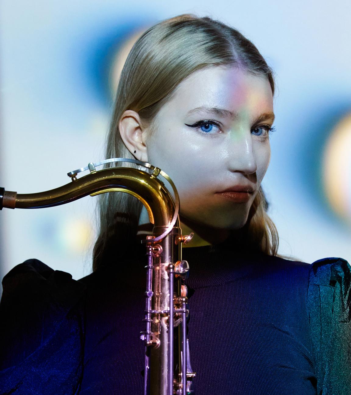 Die junge Musikerin Emma Rawicz, Kopf groß, seitlich. Das Sax seitlich vors Kinn haltend, der Saxbogen mit Mundstück gegen die Blickrichtung zeigend.